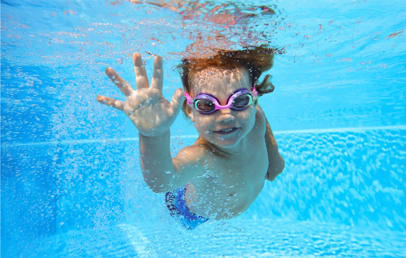 Child with purple goggles swims underwater, reaching toward the camera.