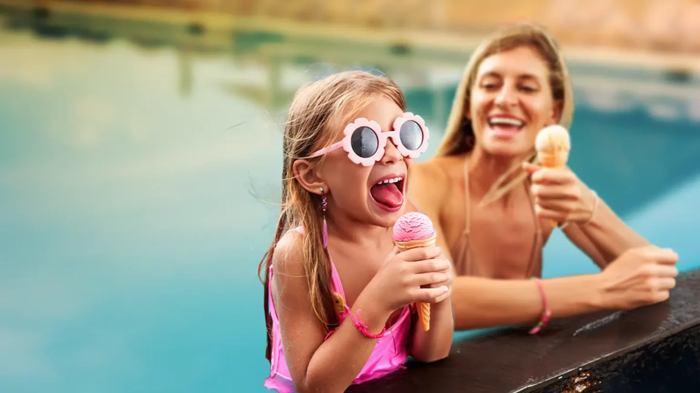 Girl with flower-shaped sunglasses and smiling woman enjoying ice cream in a pool.