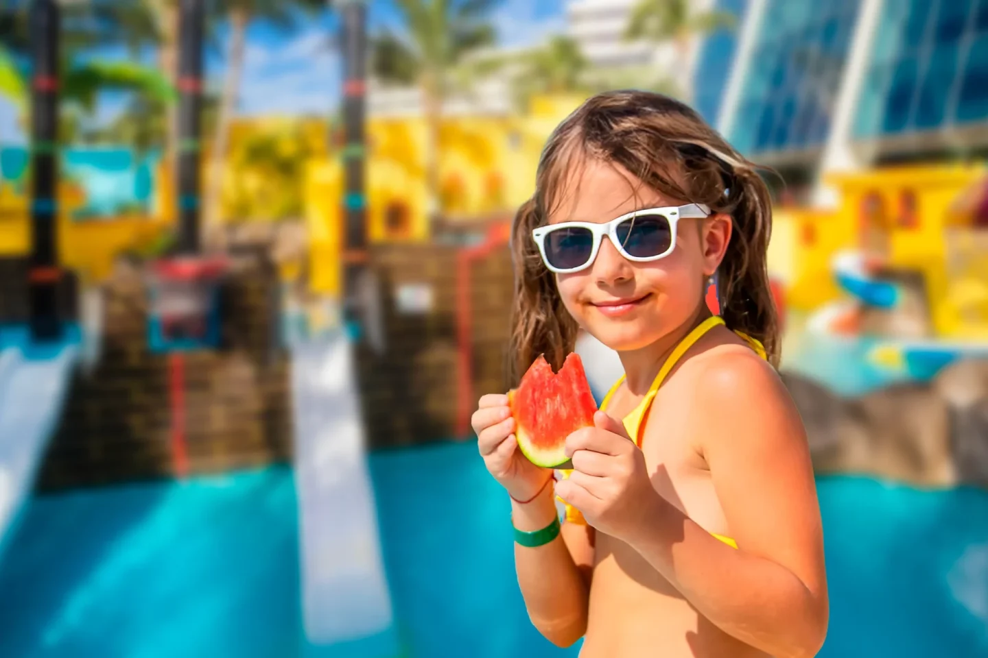 Girl in white sunglasses and yellow swimsuit holding a watermelon slice at a water park.